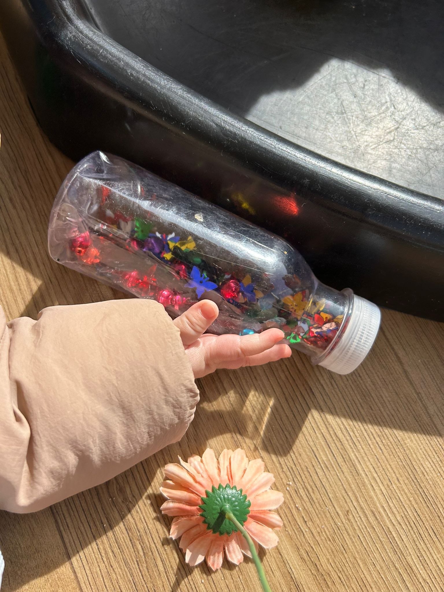 Detalle de la mano de un bebé en El Columpio explorando una botella sensorial de colores en un espacio de juego preparado.