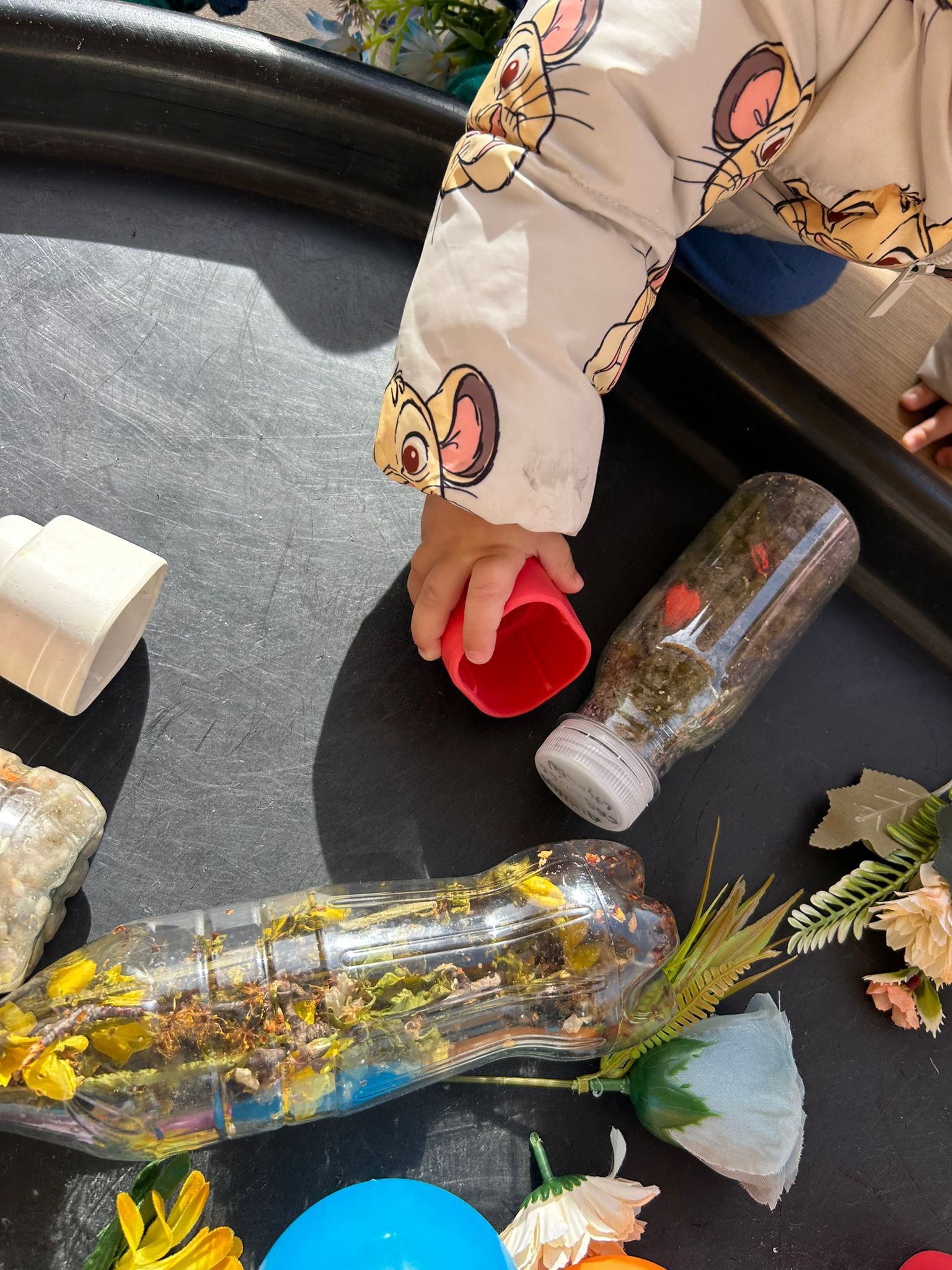 Detalle de la mano de un bebé manipulando una botella sensorial y piezas de colores en la Escuela Infantil El Columpio.