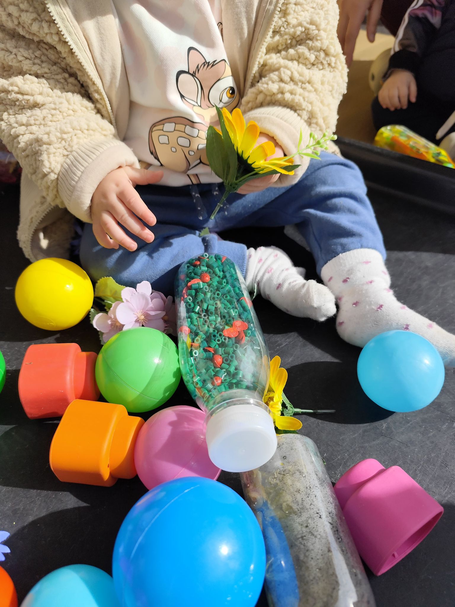 Un bebé sentado en una bandeja de juego en El Columpio, rodeado de botellas sensoriales de colores, pelotas y flores, disfrutando de una actividad multisensorial.