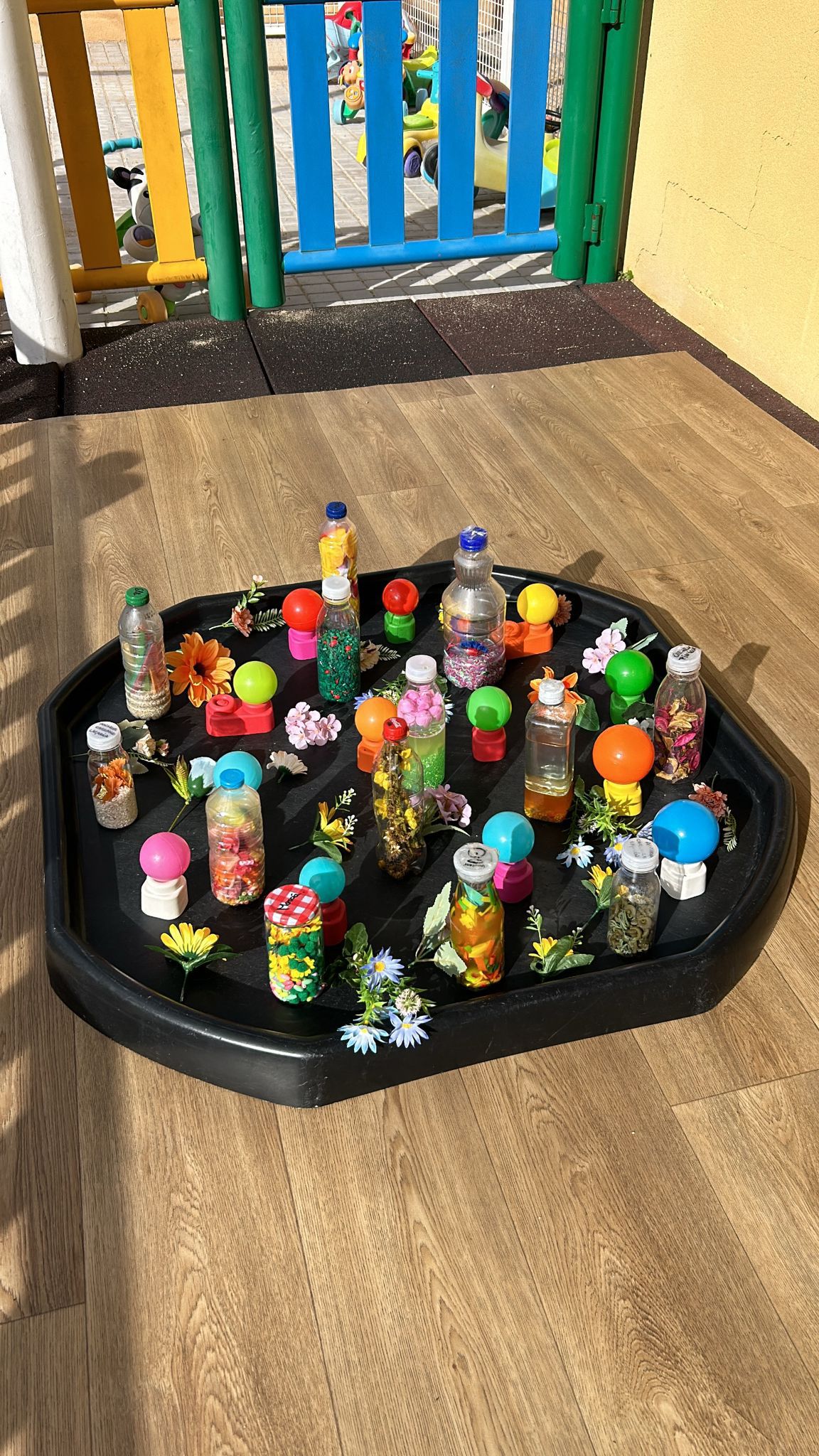 Vista general de una bandeja de experimentación con botellas sensoriales, flores y objetos de colores en el patio de la escuela infantil El Columpio.